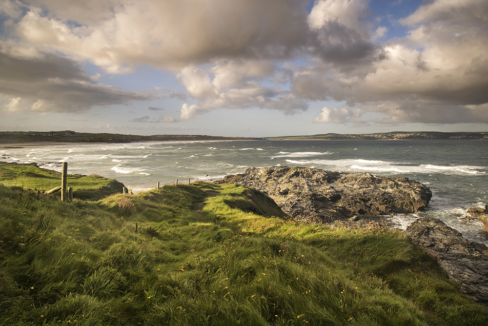 A beautiful shot of a bay taken from the vantage point of a high grassy dune overlooking the bay as the sun shines across the beach and the blue sky illuminates the surf